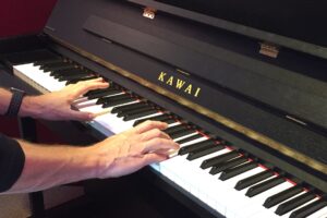 Auf diesem Bild sieht man ein akustisches Klavier der Marke Kawai das von zwei Männerhänden gespielt wird.