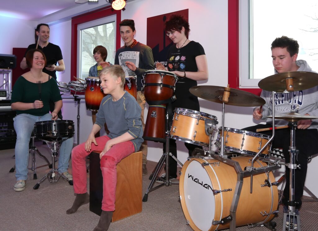 Dieses Bild zeigt 7 Personen die unterschiedliche Percussion Instrumente spielen Cajon, Djembe, Congas, Snare Drum, Schlagzeug, Cowbell
