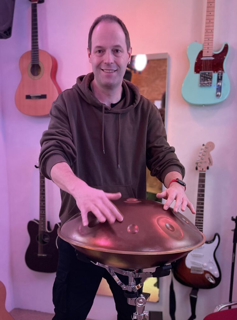 Im Vordergrund ist eine Handpan zu sehen, die von einem Mann gespielt wird. Im Hintergrund sieht man noch akustische und elektrische Gitarren